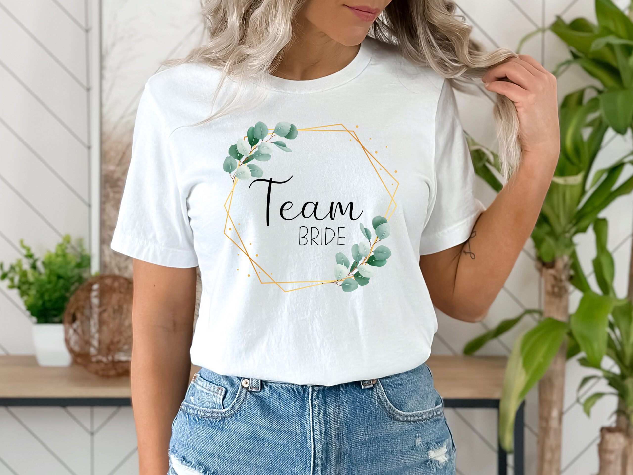 Frau trägt ein weißes T-Shirt mit dem Wort 'Team Bride' in schwarzer Schrift, umgeben von einem goldenen dünnen Rahmen und Eukalyptusblättern.