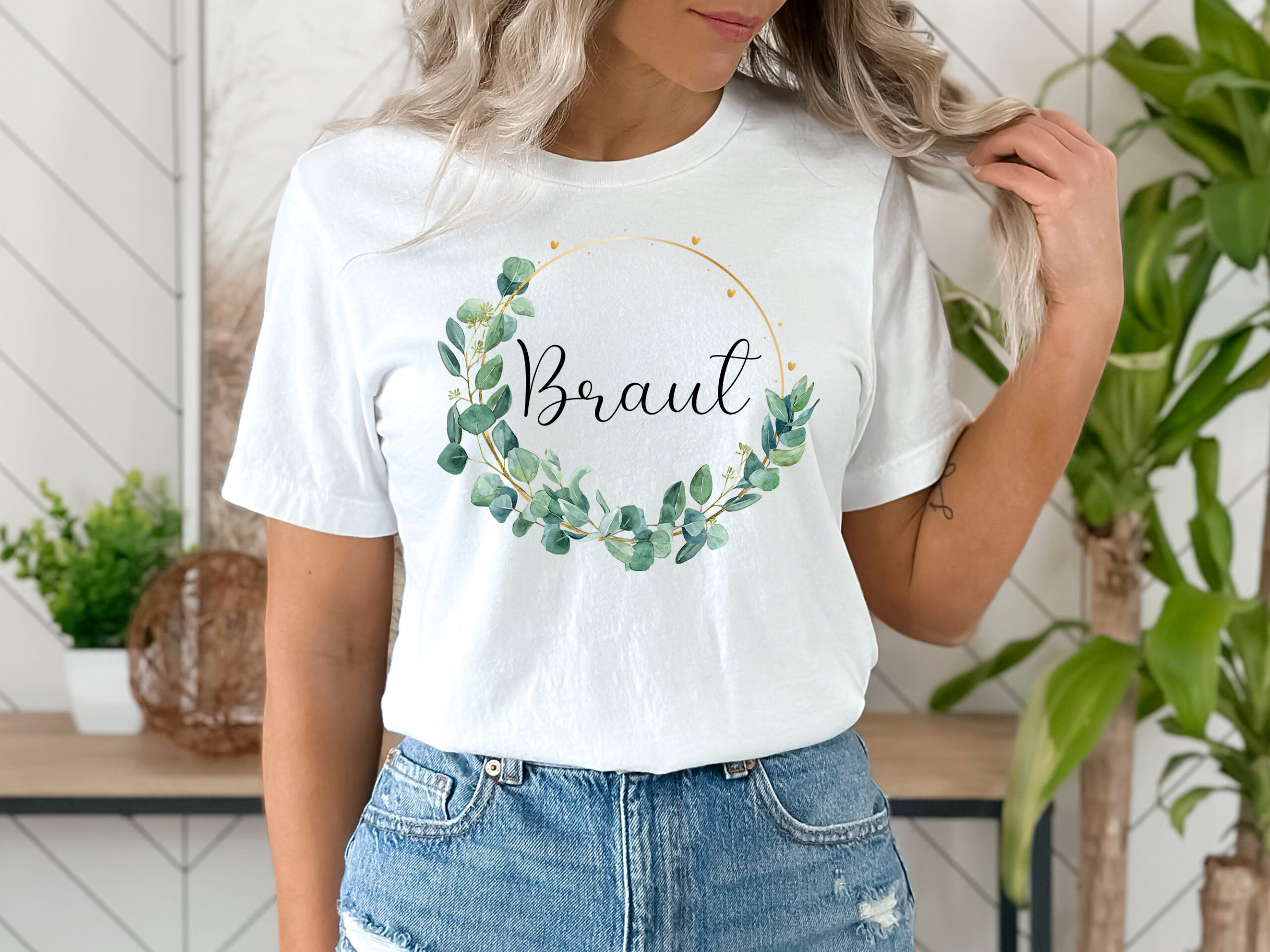 Frau trägt ein weißes T-Shirt mit dem Wort 'Braut' in schwarzer Schrift, umgeben von einem goldenen dünnen Rahmen und Eukalyptusblättern.