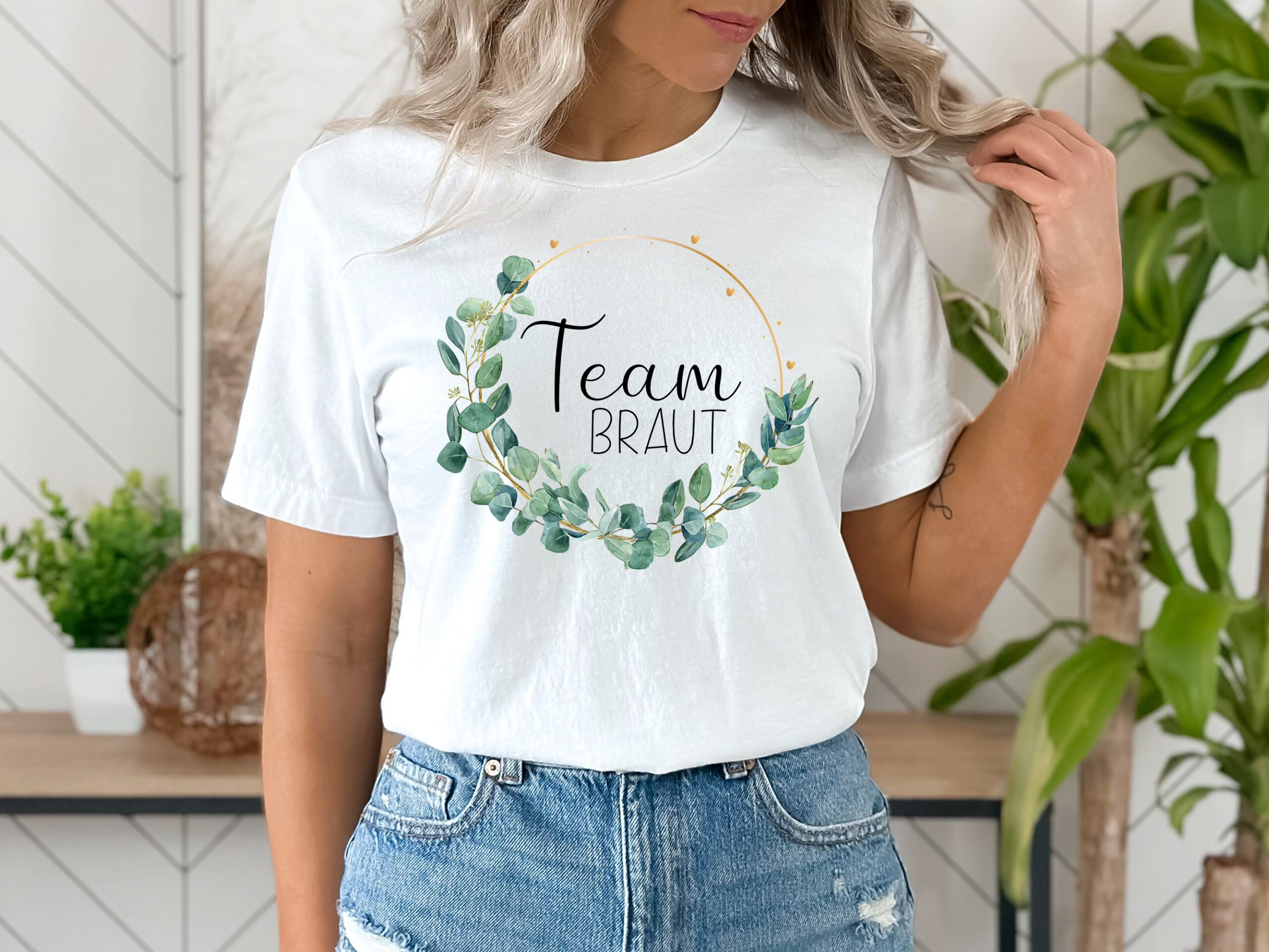 Frau trägt ein weißes T-Shirt mit dem Wort 'Team Braut' in schwarzer Schrift, umgeben von einem goldenen dünnen Rahmen und Eukalyptusblättern.