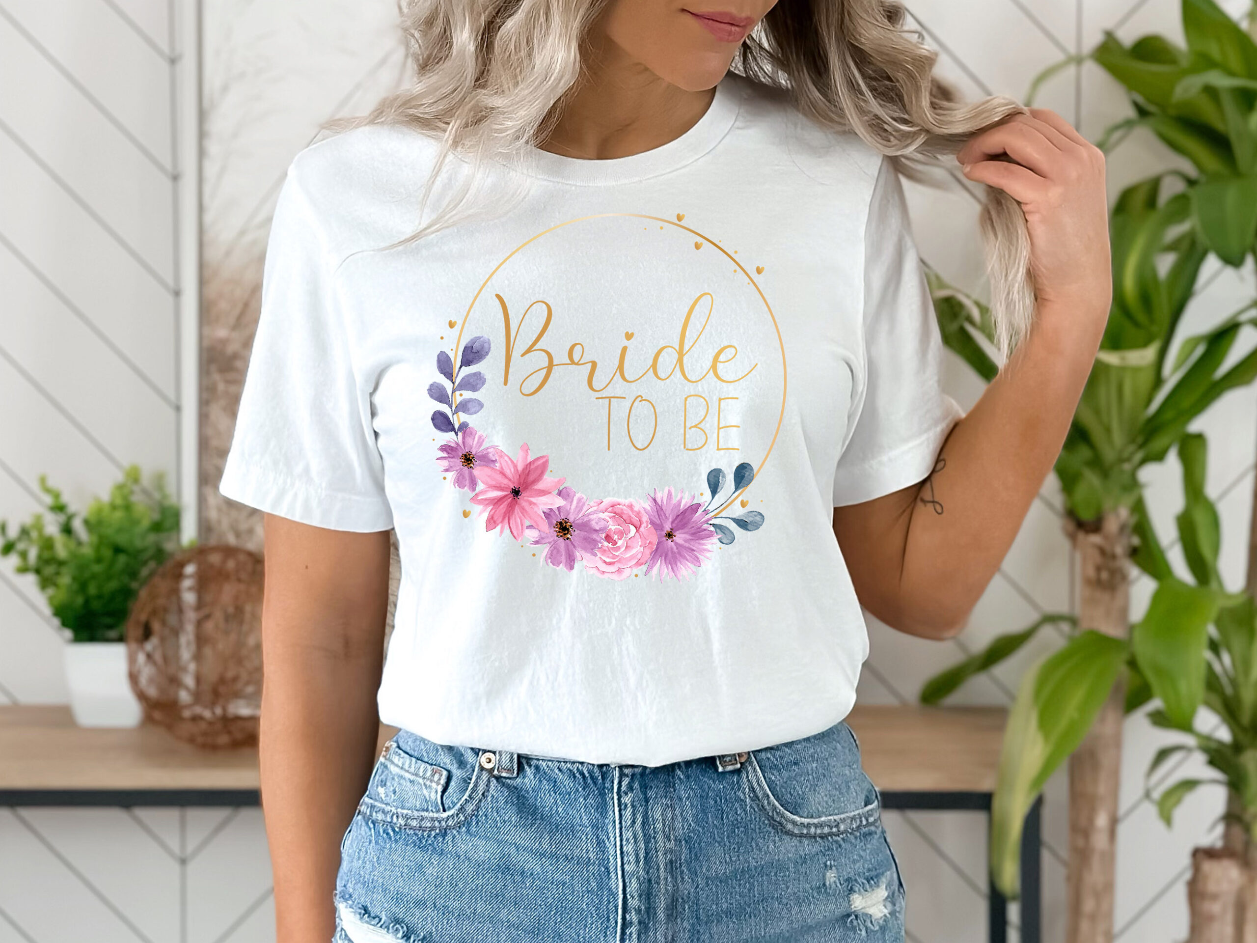 Frau trägt ein weißes T-Shirt mit dem Wort 'Bride to be' in goldener Schrift, umgeben von einem goldenen dünnen Rahmen und Blüten.